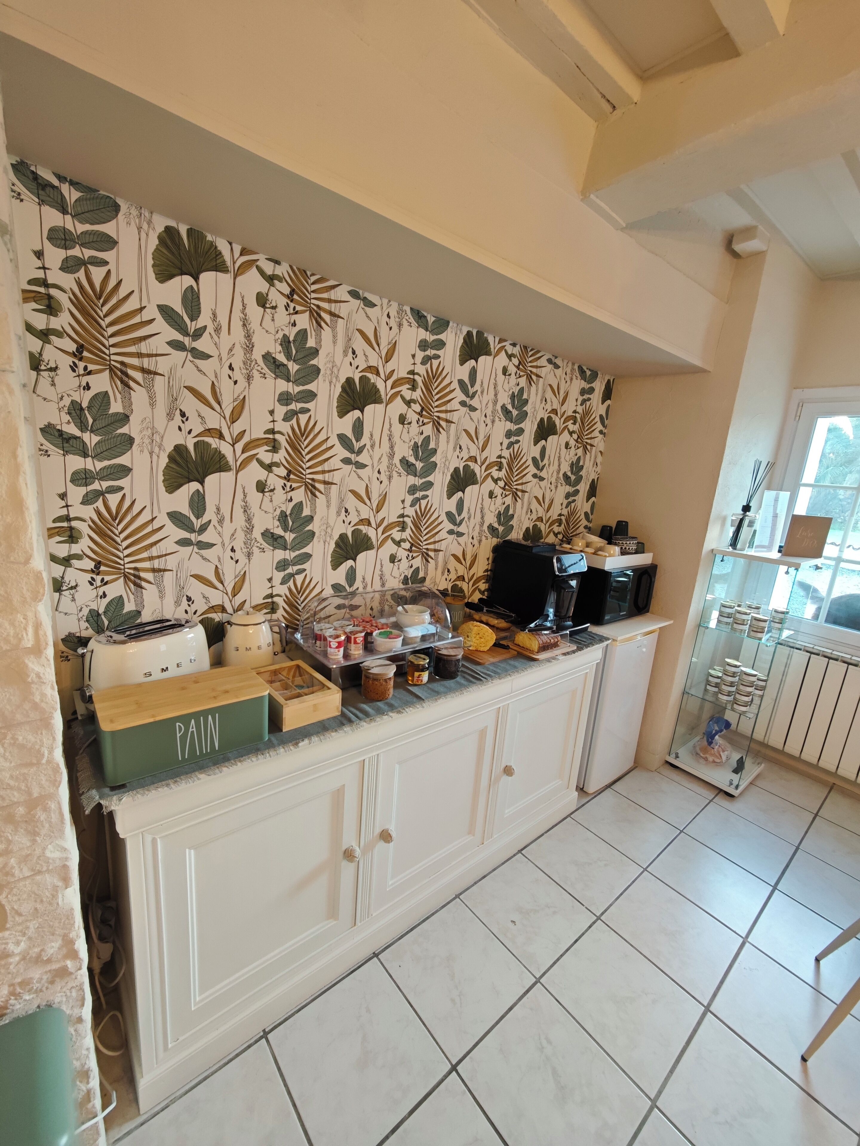 A kitchen with a patterned wallpaper, a countertop with various food items, a coffee maker, and a glass cabinet with dishes.
