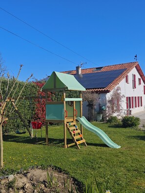 A playground with a slide and climbing structure, surrounded by a grassy yard and a house with a red-tiled roof. - La forge de Clermont - Chambres d'Hôtes (Clermont)