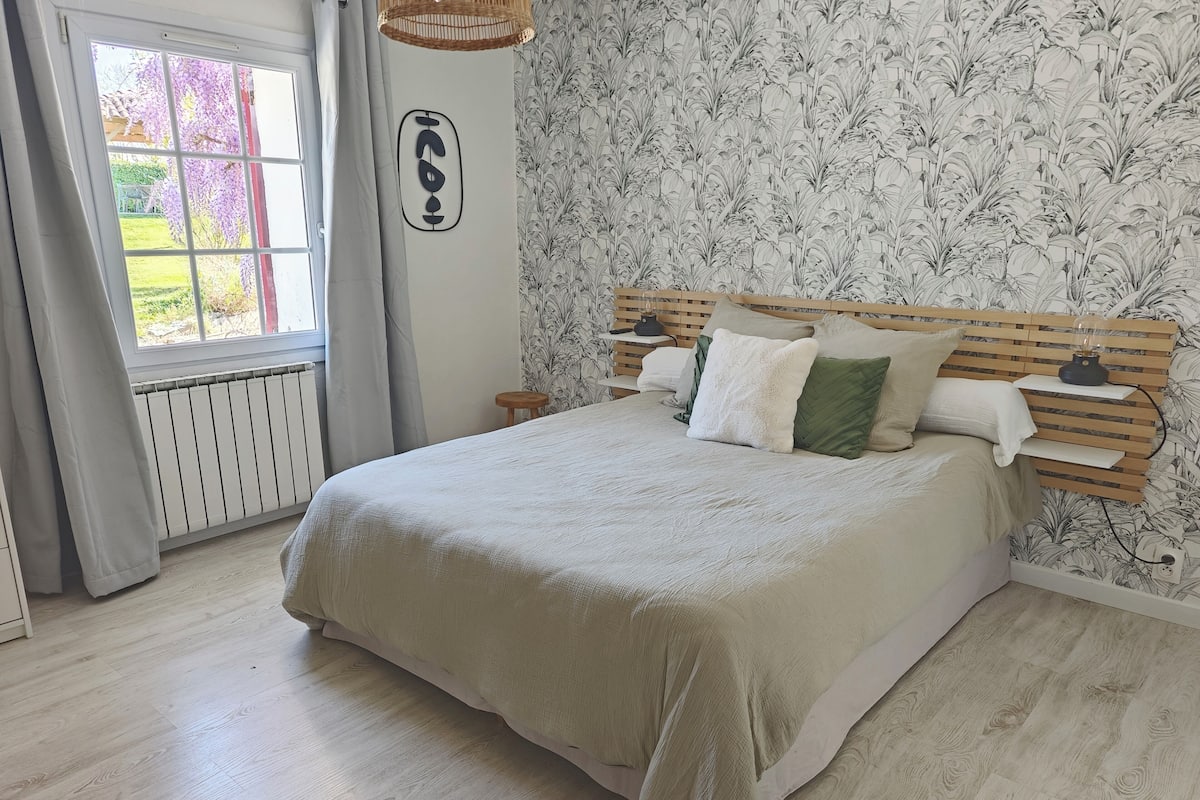 A bedroom with a bed, a nightstand, a lamp, a window with curtains, and a wall with a floral pattern.