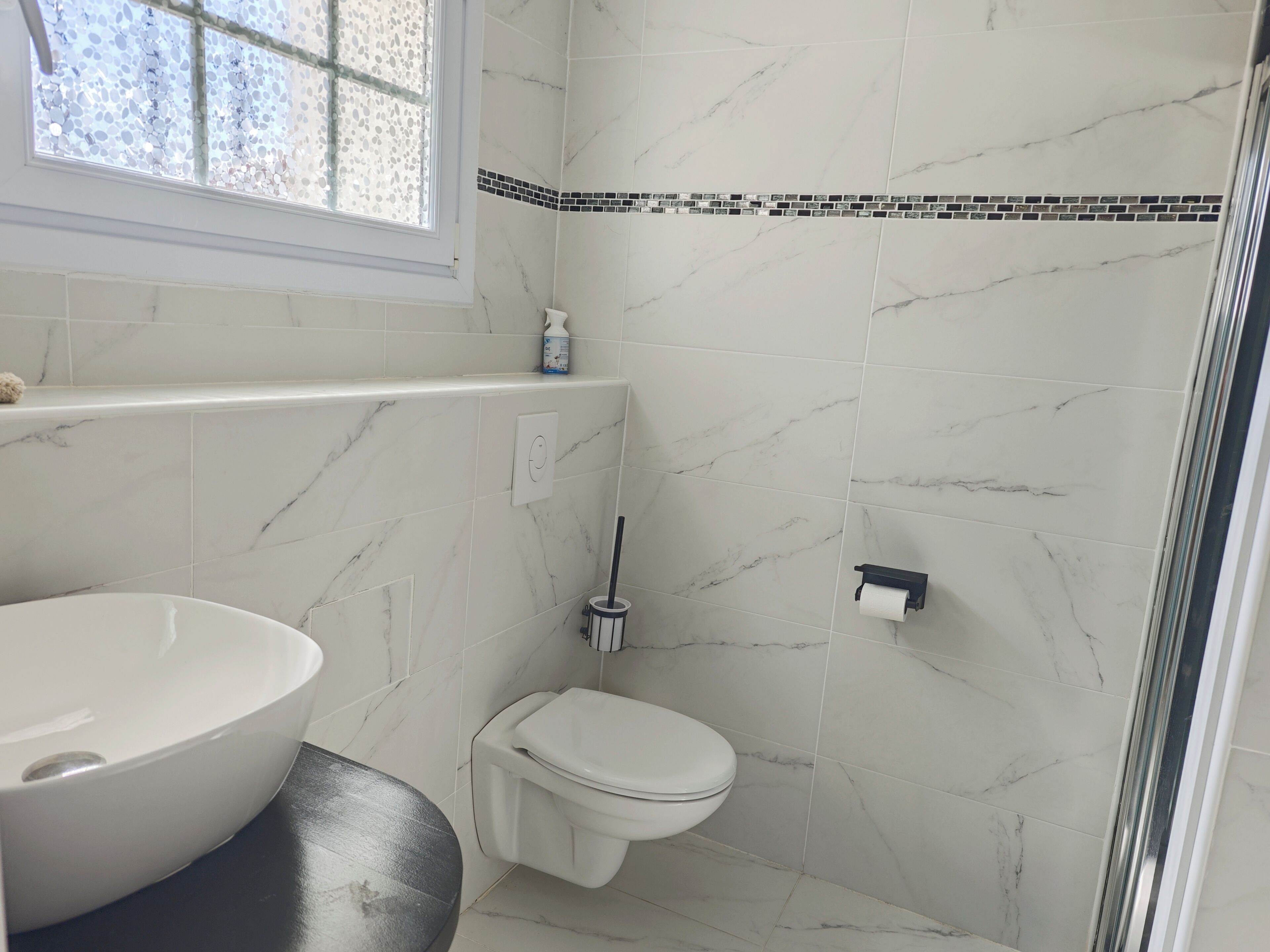 Een badkamer met een grote witte wastafel, een toilet, een douchegedeelte en een raam met uitzicht op bomen.