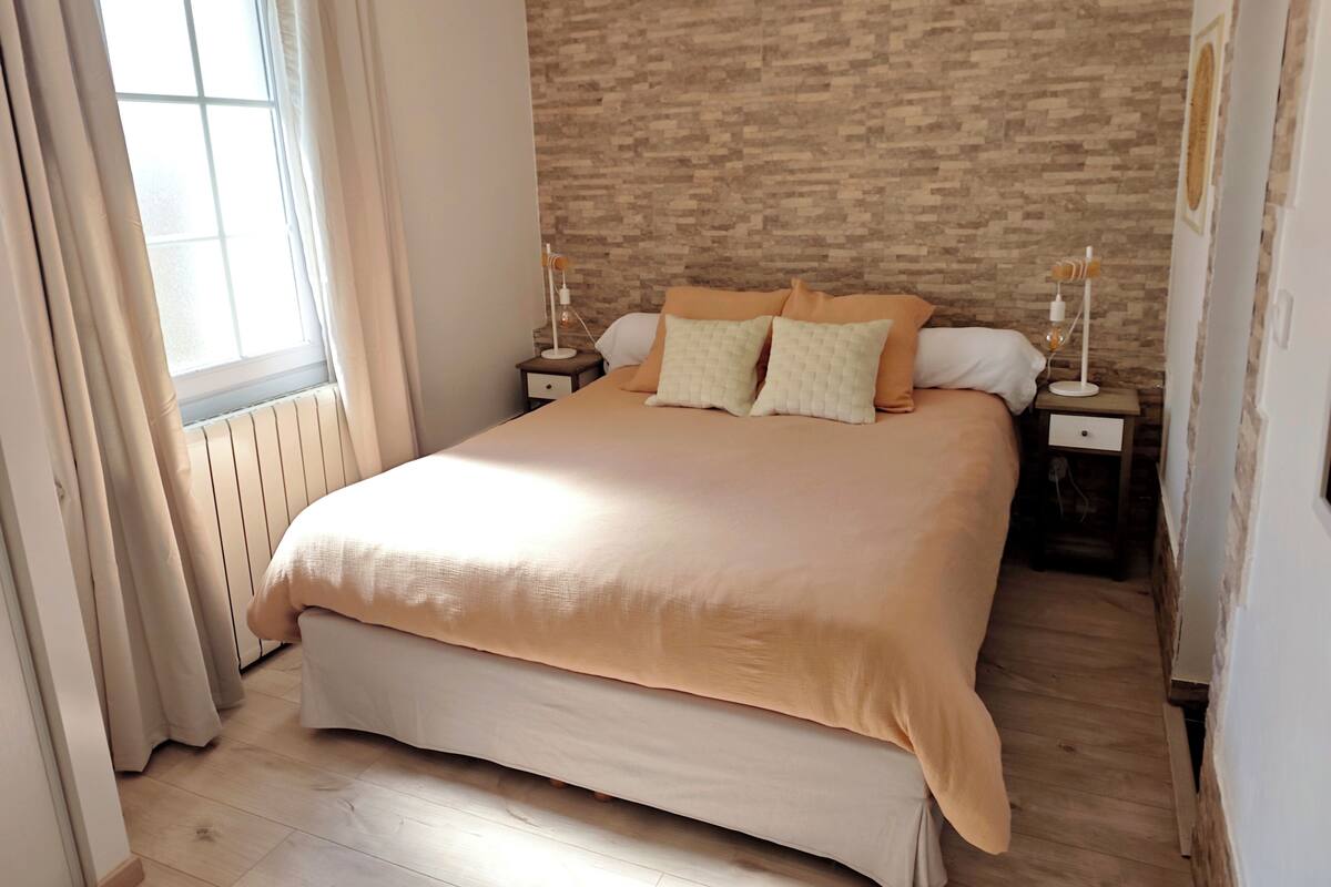 A bedroom with a large bed, two bedside tables, a window with curtains, and a brick-patterned wall.