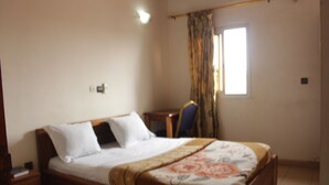 A bedroom with a bed, a nightstand, a chair, a window with curtains, and a wall-mounted light. - Hotel Massoh (Bertoua)