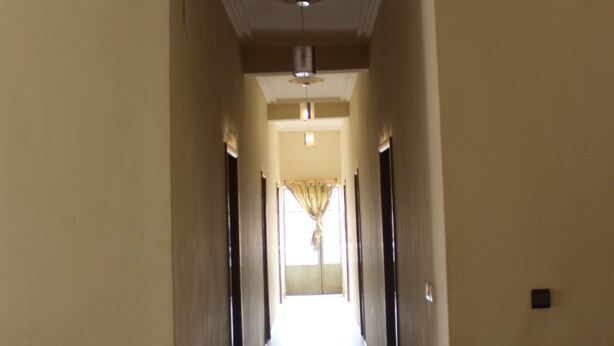 A long, narrow hallway with cream-colored walls and a tiled floor. There are doors on both sides, and a light fixture is visible on the ceiling. - Hotel Massoh (Bertoua)