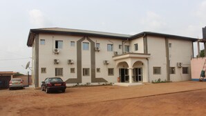 A two-story building with a flat roof, multiple windows, and a central entrance with a doorway. There are two cars parked in front. - Hotel Massoh (Bertoua)
