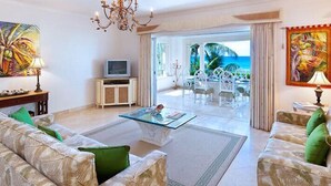 A spacious living room with a glass coffee table, two sofas with green cushions, a television on a wooden cabinet, and a view of the ocean through large windows. - Schooner Bay 201 - Condo Flamboyant (Speightstown)