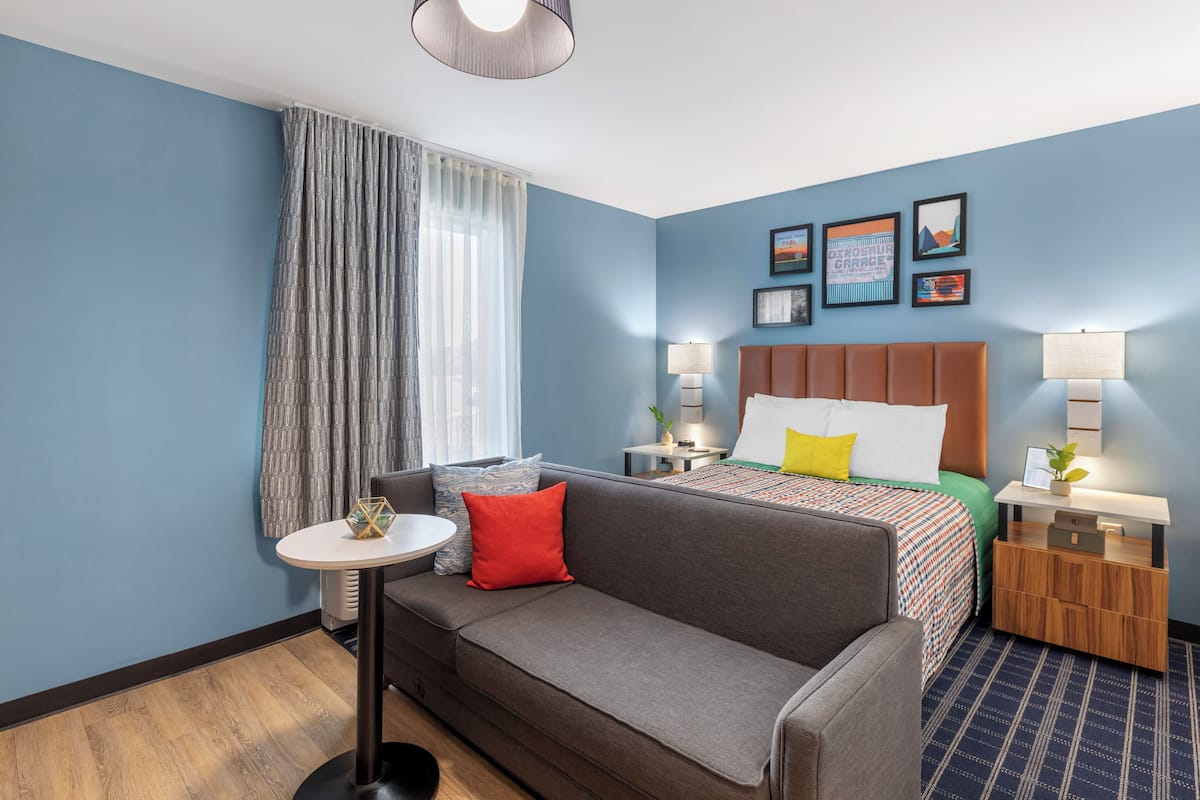 A hotel room with a grey sofa, a small round table, a bed with a colorful bedspread, and a wooden nightstand with a lamp.
