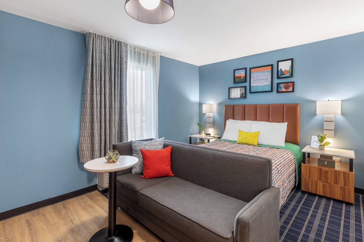 Een hotelkamer met een grijze bank, een klein rond tafeltje, een bed met een kleurrijk beddegoed en een houten nachtkastje met een lamp.