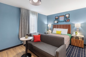 A hotel room with a grey sofa, a small round table, a bed with a colorful bedspread, and a wooden nightstand with a lamp. - Uptown Suites Extended Stay Denver CO – Centennial (Centennial)