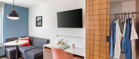 A modern hotel room with a grey sofa, a small round table, a desk with a chair, a TV mounted on the wall, and a wooden wardrobe with clothes hanging.