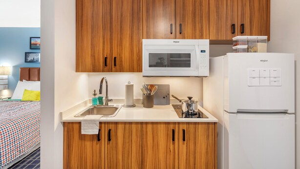 A compact kitchen with wooden cabinets, a microwave, a refrigerator, and a sink. - Uptown Suites Extended Stay Tampa FL – Riverview (Riverview)