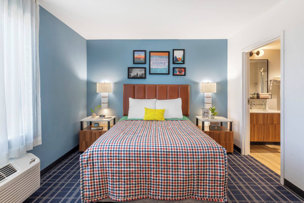 A hotel room with a checkered bedspread, a headboard, bedside lamps, a nightstand, a window with curtains, and a wall with framed pictures.