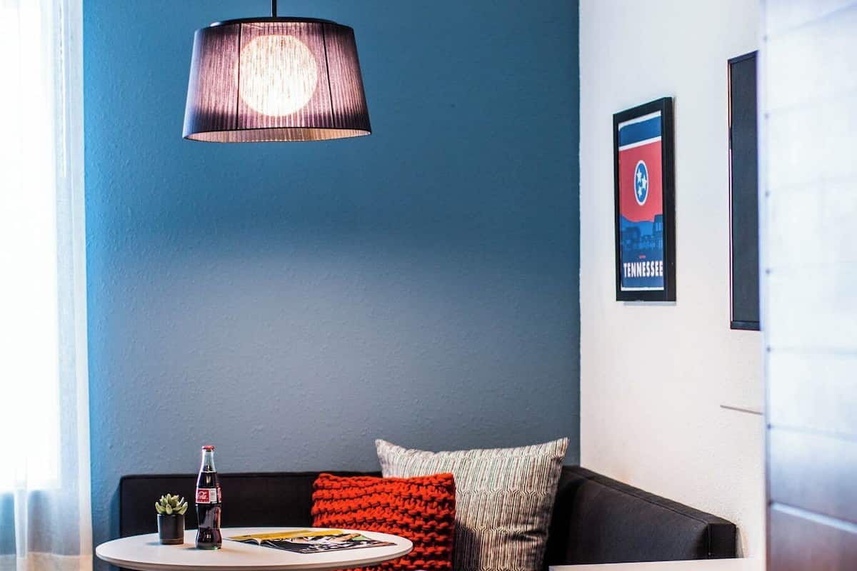 A modern room with a blue wall, a round table with a bottle, a sofa with a red cushion, a pink chair, and a hanging light fixture.