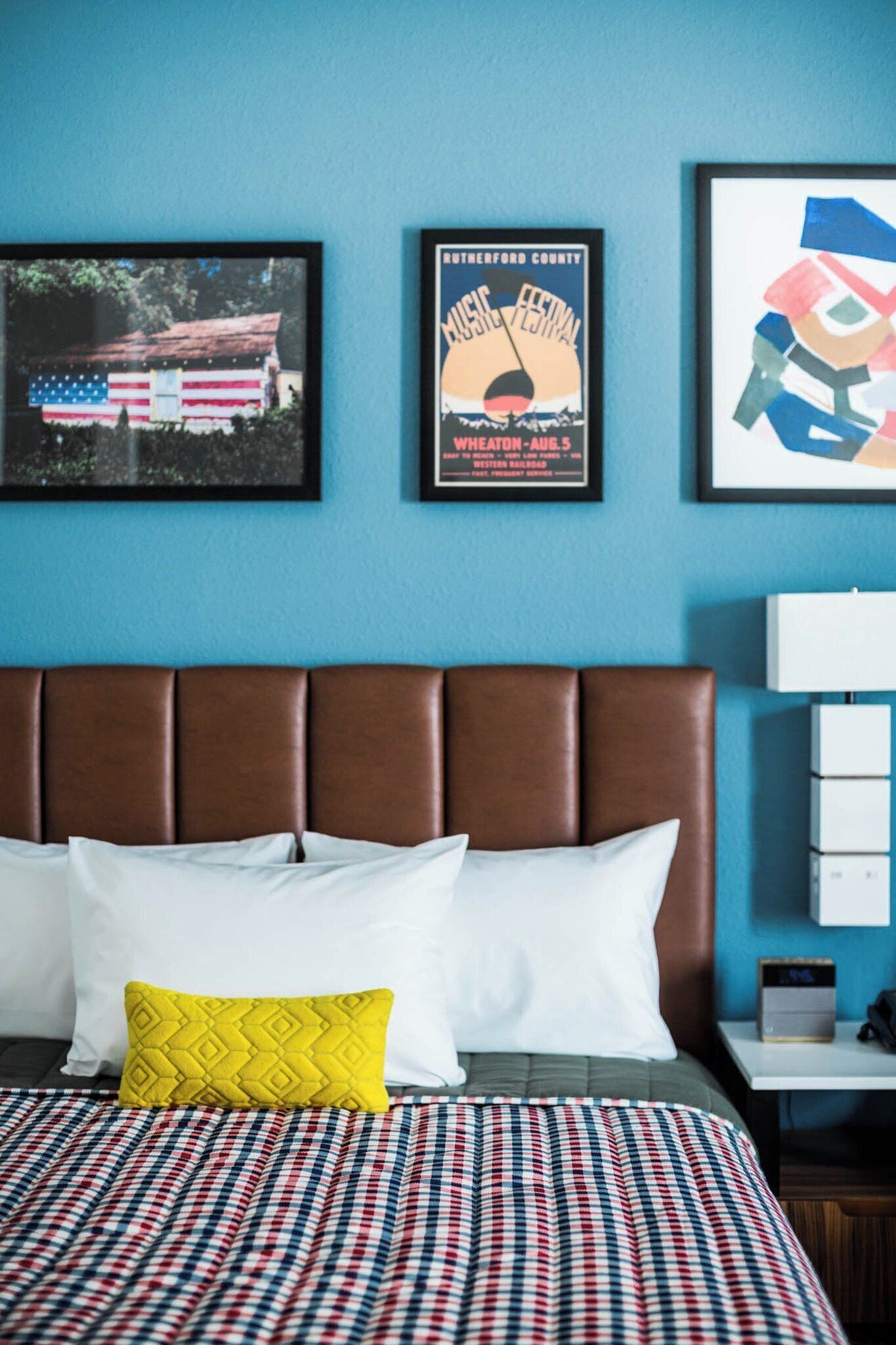 A hotel room with a bed, a headboard, a nightstand, and wall art including a poster and a framed picture.
