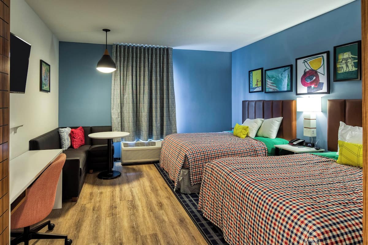 Ein Hotelzimmer mit zwei Betten, einem Schreibtisch, einem Stuhl, einem kleinen Tisch, einer Lampe und einem Fenster mit Vorhängen.