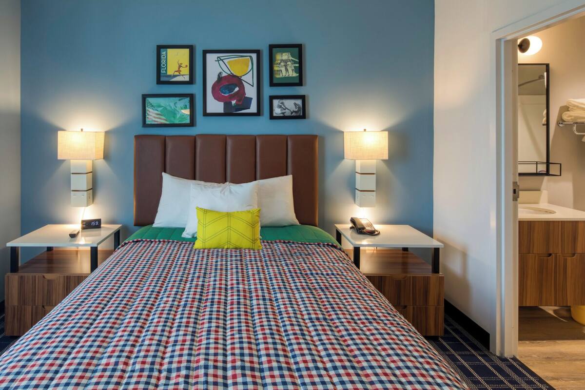 Ein Hotelzimmer mit kariertem Bettbezug, braunem Kopfteil, zwei Nachttischen mit Lampen, einem Beistelltischchen mit Telefon und Bildern an der Wand.