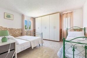 A bedroom with two beds, a wardrobe, a bedside table with a lamp, a window with curtains, and a stone wall. - 1136 Villetta Paola by Barbarhouse (Melendugno)
