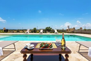 A poolside table with a tray of watermelon slices, a bottle of champagne, and glasses, overlooking a clear blue pool with lounge chairs and a stone wall in the background. - 1136 Villetta Paola by Barbarhouse (Melendugno)