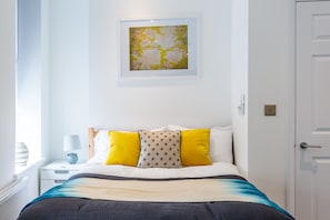 Quarto arrumado com uma cama, almofadas, mesa de cabeceira com abajur e uma obra de arte emoldurada na parede.