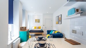 A modern kitchen with a dining area, a bed with blue and yellow bedding, and a blue sofa. - Blue Suede Shoes (Southend-on-Sea)