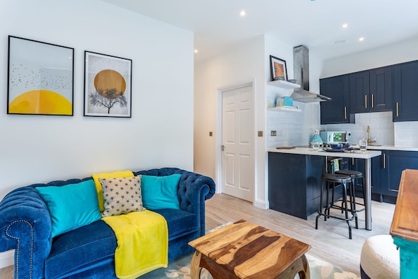 Uma sala de estar moderna com um sofá azul, uma almofada amarela e uma mesa de centro de madeira. Próximo, encontra-se uma cozinha com armários escuros, eletrodomésticos de aço inoxidável e uma pequena ilha com bancos.