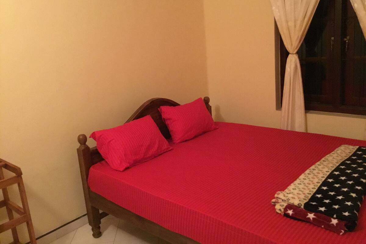 Una cama con colcha y almohadas rojas, cabecera de madera y ventana con cortinas.