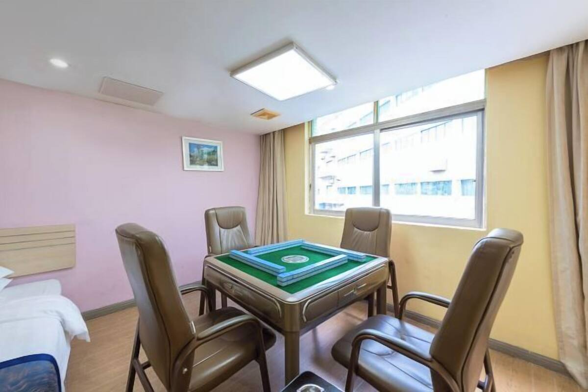 Habitación de hotel con una mesa de billar verde, cuatro sillones de cuero, una cama con ropa blanca, un pequeño cuadro en la pared y una ventana con cortinas.