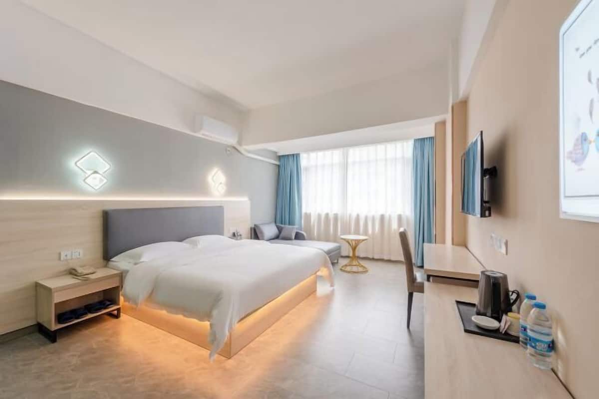 A modern hotel room with a neatly made bed, a bedside table with a lamp, a small desk with a chair, and a large window with curtains.