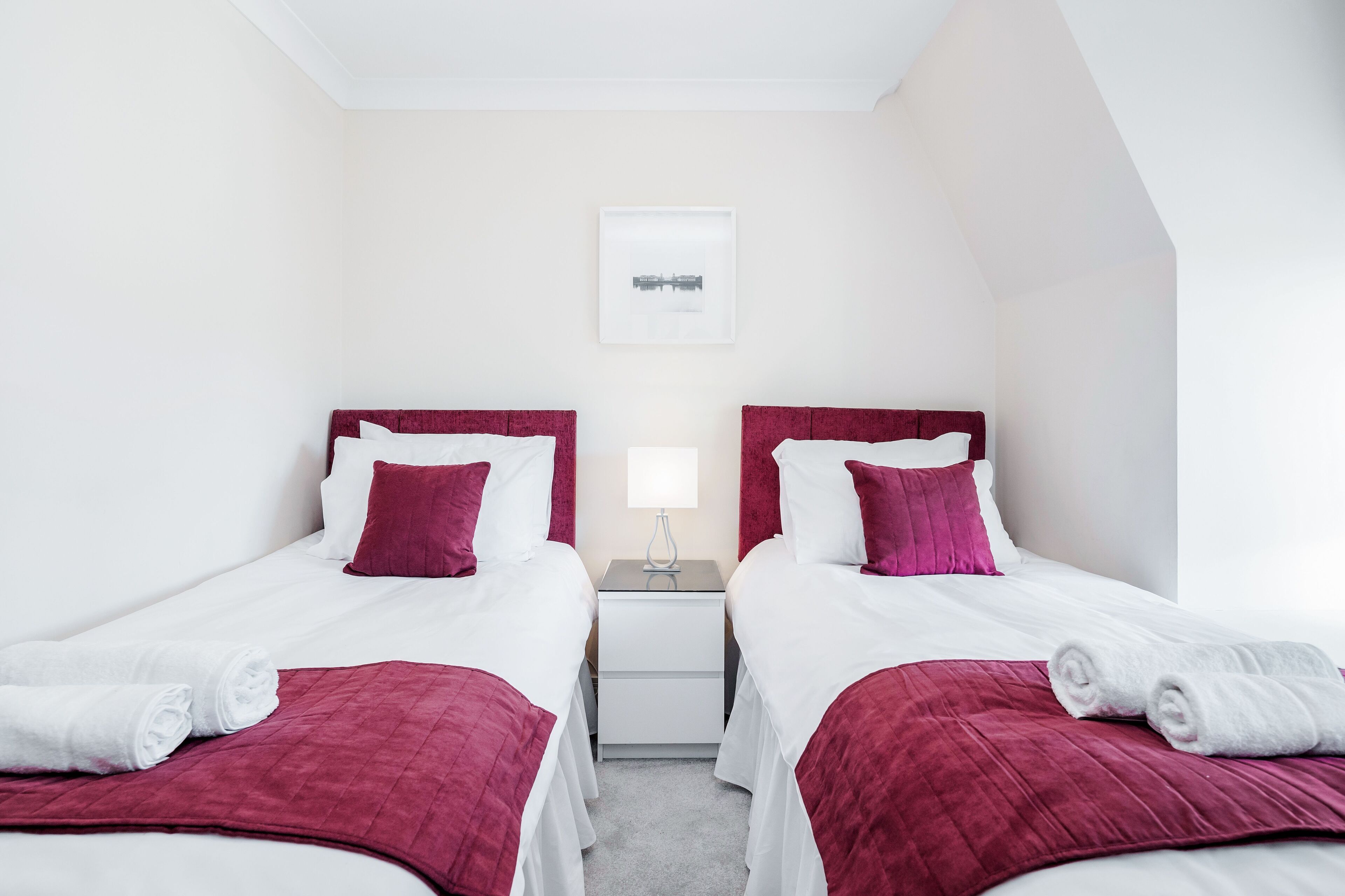 A hotel room with two beds, each with white bedding and red headboards, a nightstand with a lamp, and a framed picture on the wall.