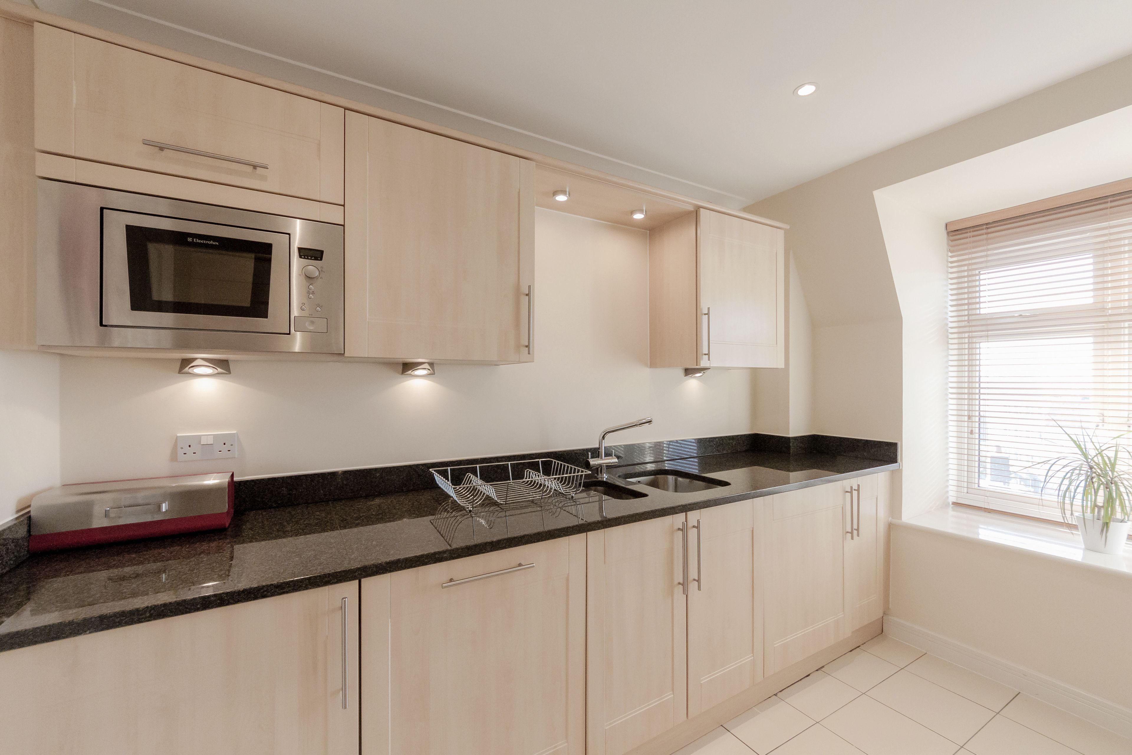 A modern kitchen with light wood cabinets, a stainless steel microwave, and a black countertop.