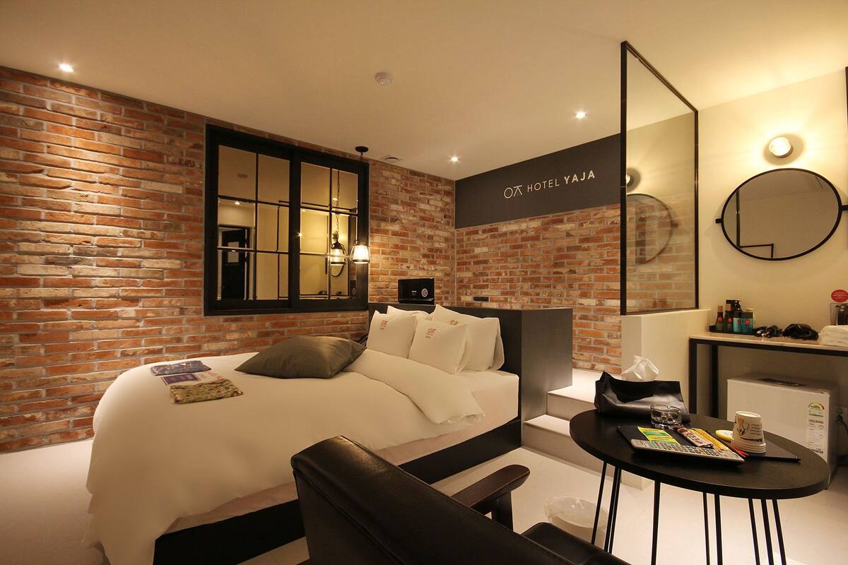 Una habitación de hotel moderna con pared de ladrillo, una cama grande, un escritorio con silla y un espejo redondo.