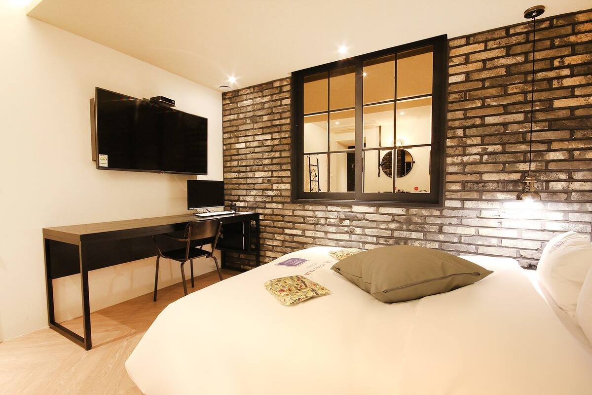 Un dormitorio moderno con pared de ladrillo, cama con almohadas, escritorio con silla y televisión montada en la pared.