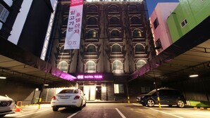 L’entrée d’un hôtel, avec une façade moderne, un éclairage décoratif et un parking où sont garées des voitures.