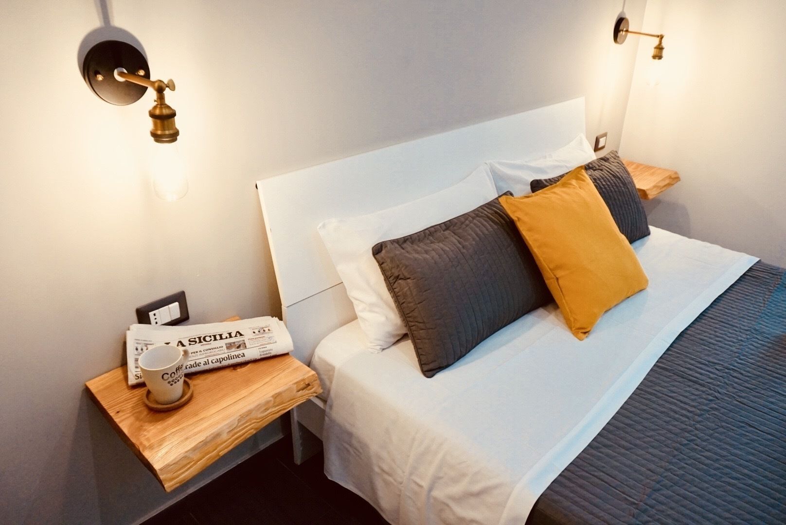 Un lit bien fait, avec des oreillers blancs et foncés, une table de chevet en bois sur laquelle reposent un journal et une tasse de café, et des lampes fixées au mur.