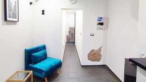 A narrow hallway with a blue sofa, a wooden side table, a clock, and a wall-mounted air conditioner. - Passi di Mare Rooms (Giardini Naxos)