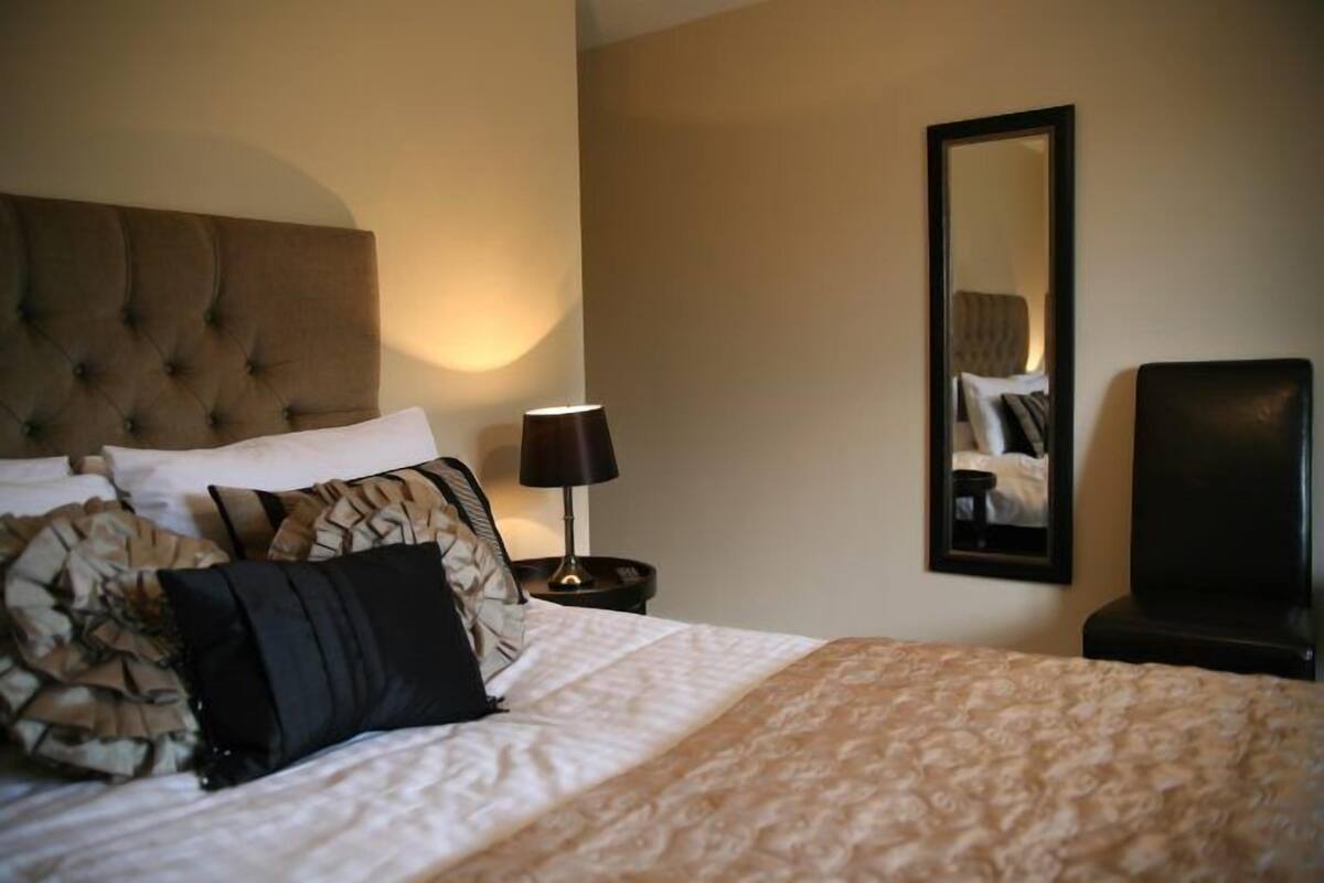 A hotel room with a large bed, a headboard, a bedside table with a lamp, a mirror, and a chair.