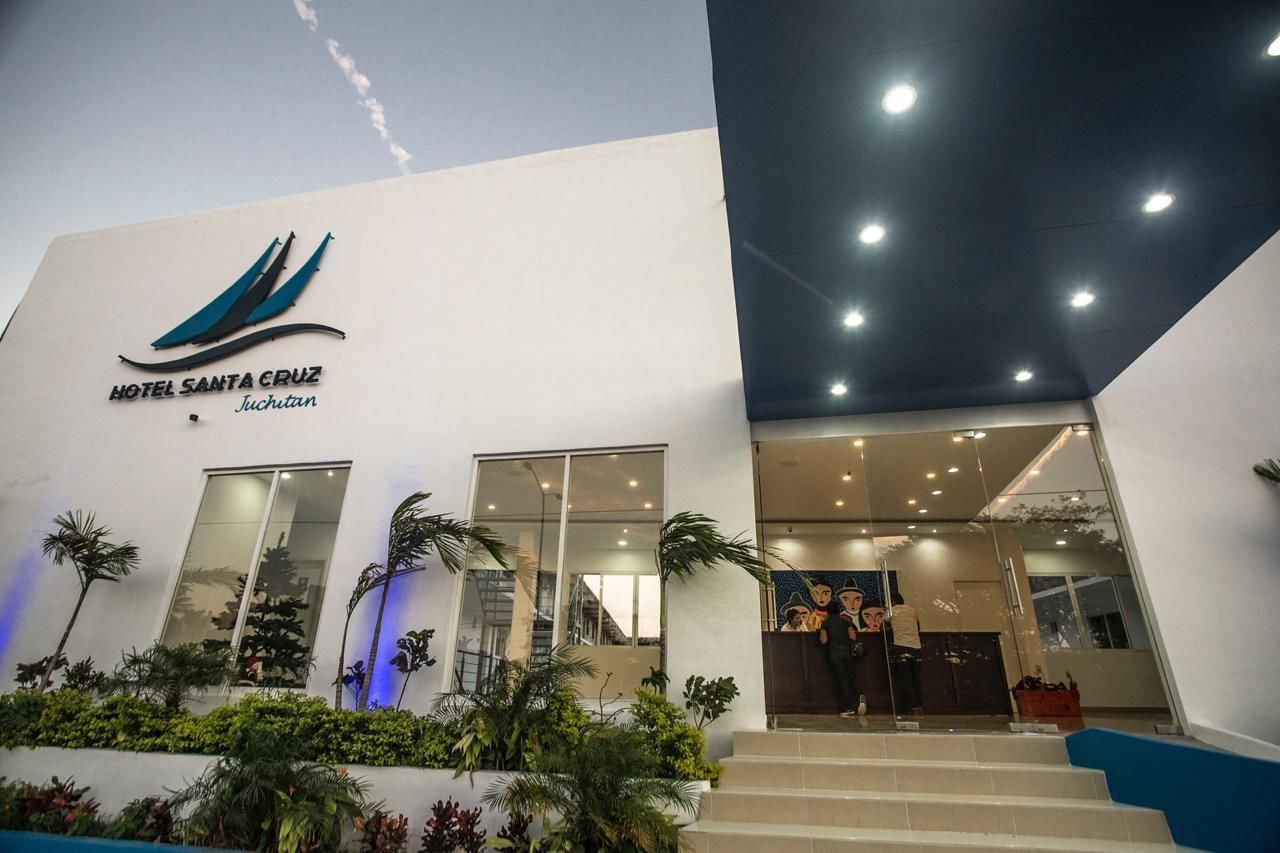 The exterior of Hotel Santa Cruz in Juchitan, featuring a modern design with a prominent logo, large windows, and a well-lit entrance.