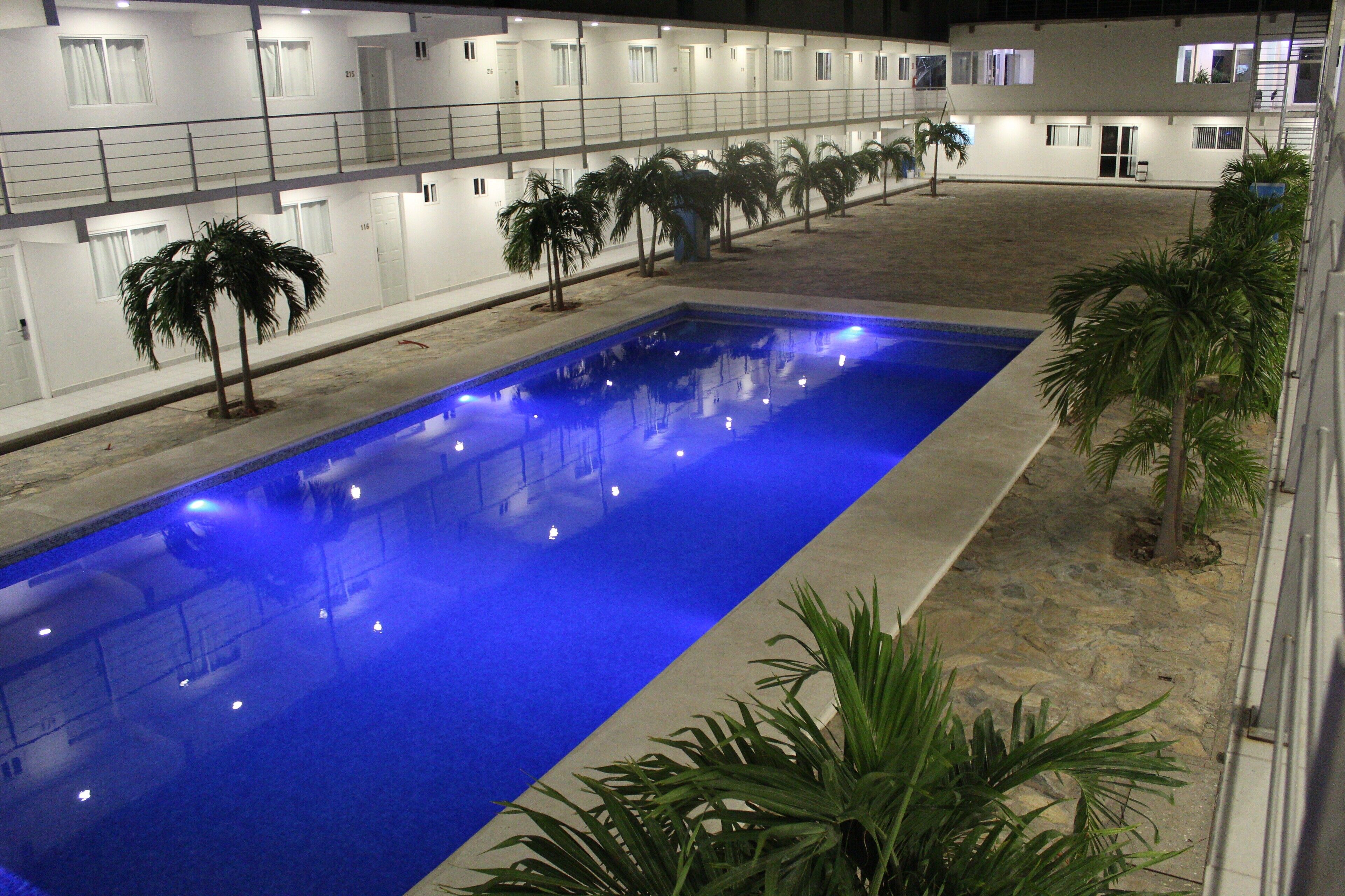 A swimming pool with blue lighting, surrounded by palm trees and a building with multiple balconies.