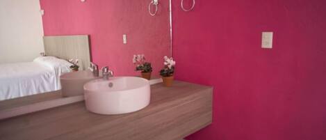 A modern bathroom with a large round sink, a bed, and a pink wall.