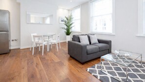 A modern living room with a grey sofa, white chairs, a glass coffee table, and a large window providing natural light. - Roomspace Apartments -Sterling House (London)