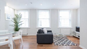A modern living room with a grey sofa, white chairs, a glass coffee table, and a large potted plant. - Roomspace Apartments -Sterling House (London)