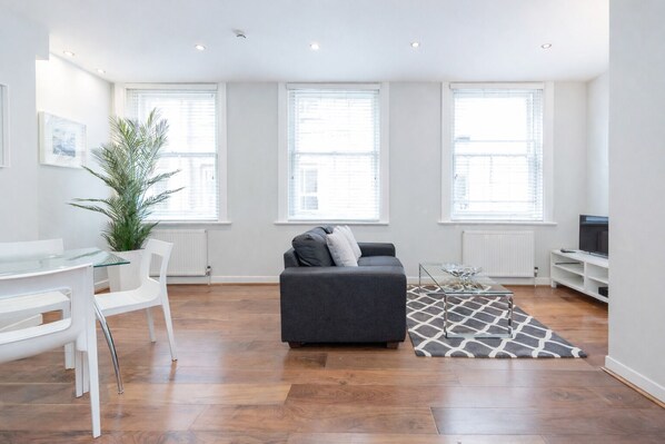 A modern living room with a grey sofa, white chairs, a glass coffee table, and a large potted plant. - Roomspace Apartments -Sterling House (London)