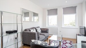 A modern living room with a grey sofa, glass shelves, a coffee table, and large windows. - Roomspace Apartments -Swan House (Leatherhead)