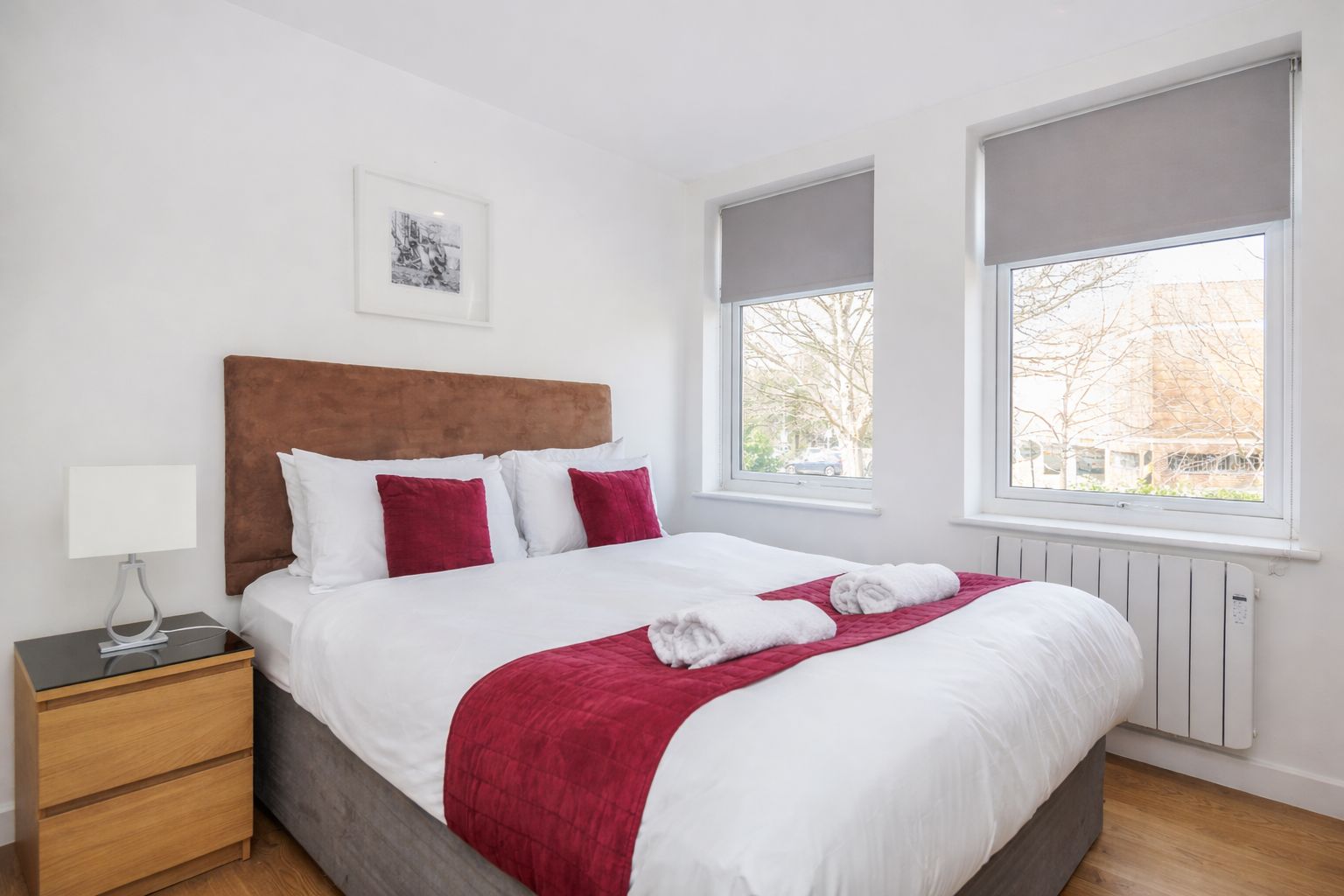 A neatly made bed with white and red bedding, a bedside table with a lamp, and a window with a view of trees and buildings.