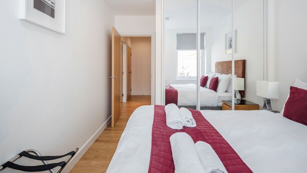 A hotel room with a large bed, a wardrobe, a bedside table, and a window. - Roomspace Apartments -Swan House (Leatherhead)