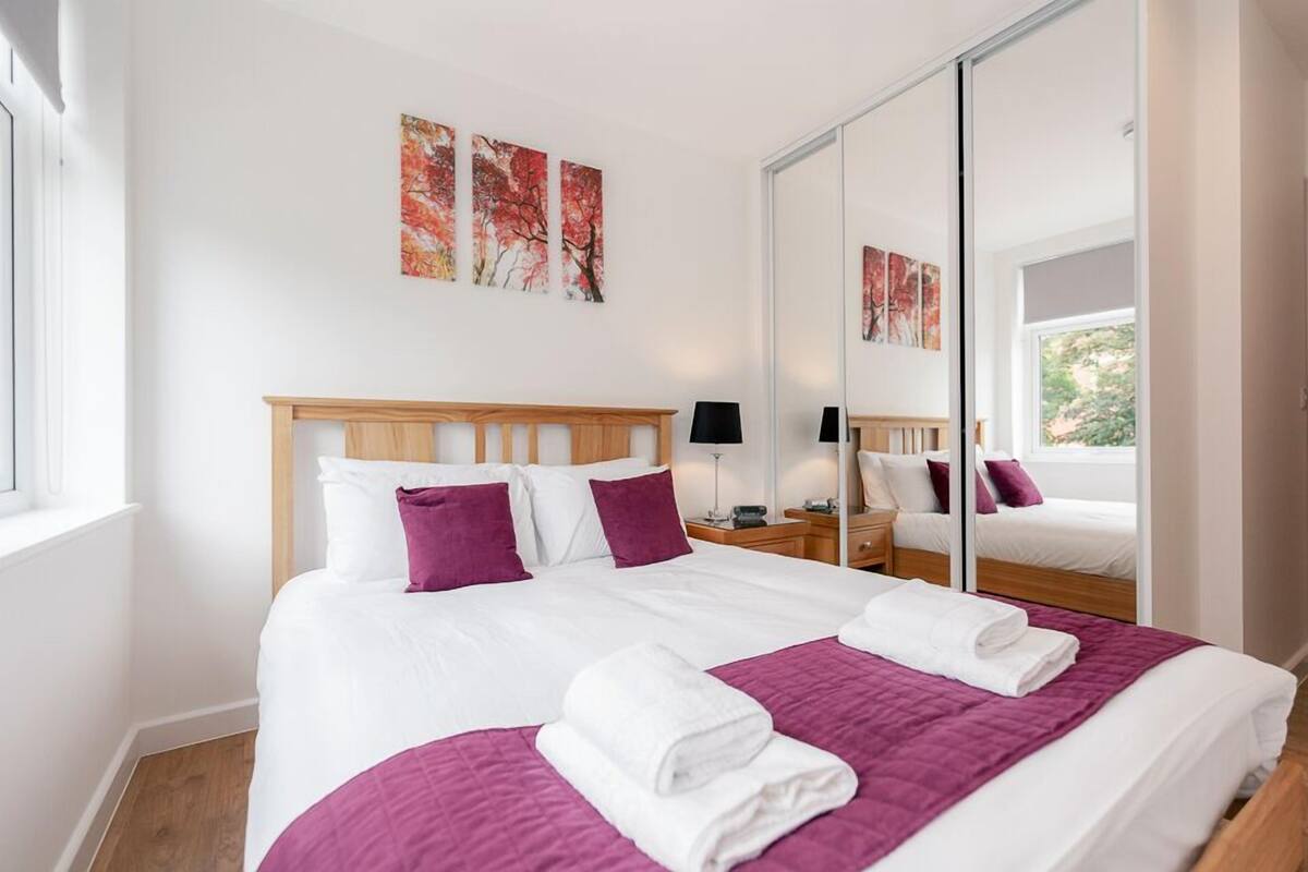 A modern hotel room with a large bed, a wooden headboard, and a large mirror. There are two paintings on the wall and a window with a view of greenery.