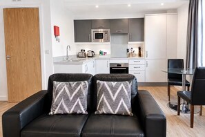 A modern living room with a black leather sofa, two patterned cushions, a glass dining table, and a kitchen in the background. - Roomspace Apartments -Swan House (Leatherhead)