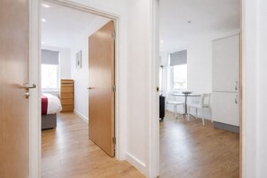 A modern interior with a wooden door, a bed with a red accent, a dining area with a glass table and white chairs, and a white wardrobe. - Roomspace Apartments -Swan House (Leatherhead)