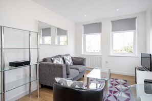 A modern living room with a grey sofa, a glass coffee table, and a TV stand. - Roomspace Apartments -Swan House (Leatherhead)