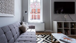A modern living room with a grey sofa, a patterned rug, a white coffee table, a bookshelf, and a large window with a view of a brick building. - Californication (Southend-on-Sea)