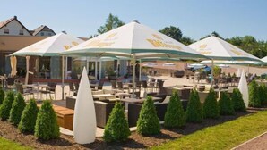 Un espace extérieur aménagé avec des parasols, des tables et des chaises, entouré de petits arbres et bordé d’un sentier.